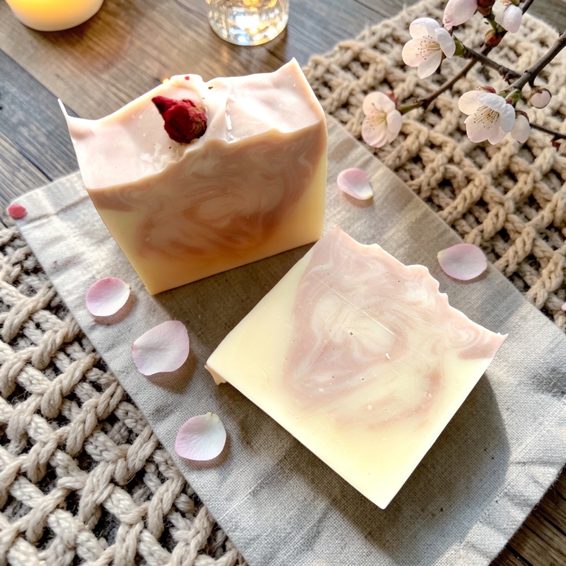 Two artisan cold-process soap bars with pink swirls and a dried rosebud on a woven mat, cherry blossom nearby.